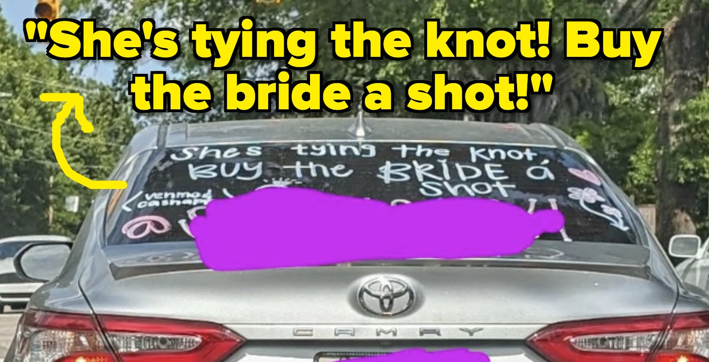 Car with "She's tying the knot! Buy the bride a shot!" written on rear window driving on street. Trees, other cars, and traffic lights are visible