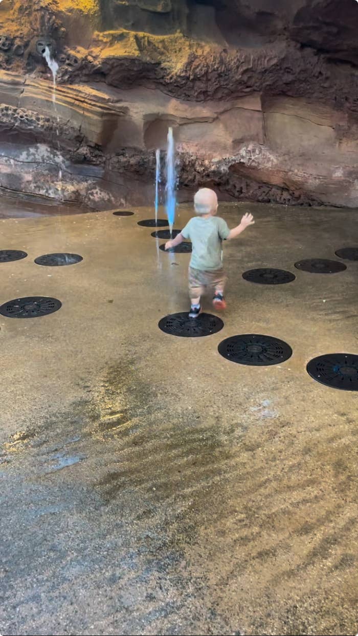 A toddler plays with a water fountain coming from the ground, surrounded by rock formations