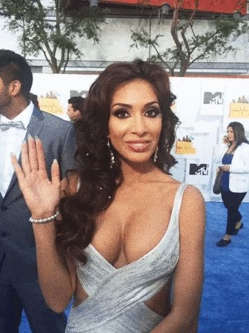 Farrah Abraham waves while posing on a blue carpet at an MTV event. She wears a plunging, sleeveless dress with cutouts, styled hair, and earrings.