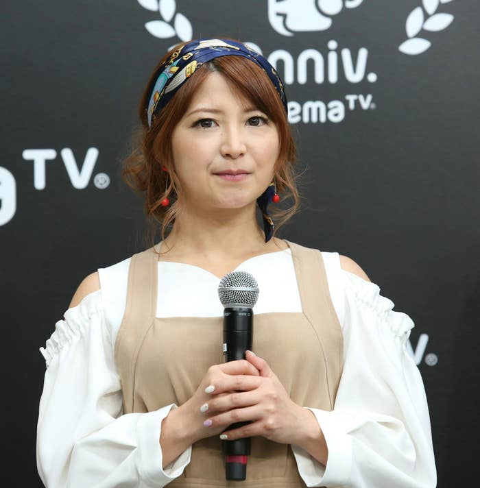 女性がインタビューに応じて話している姿。オフショルダーのシャツとエプロンスタイルのドレスを着用。背景にロゴがある。