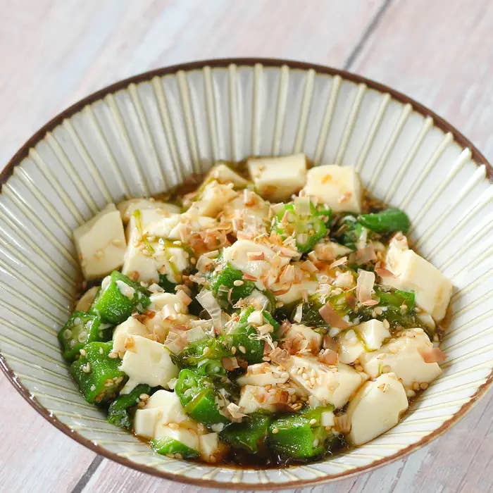 白い陶器のボウルに盛り付けられた、刻んだ豆腐とオクラの和え物。上にゴマと細かく刻んだナッツがトッピングされている。