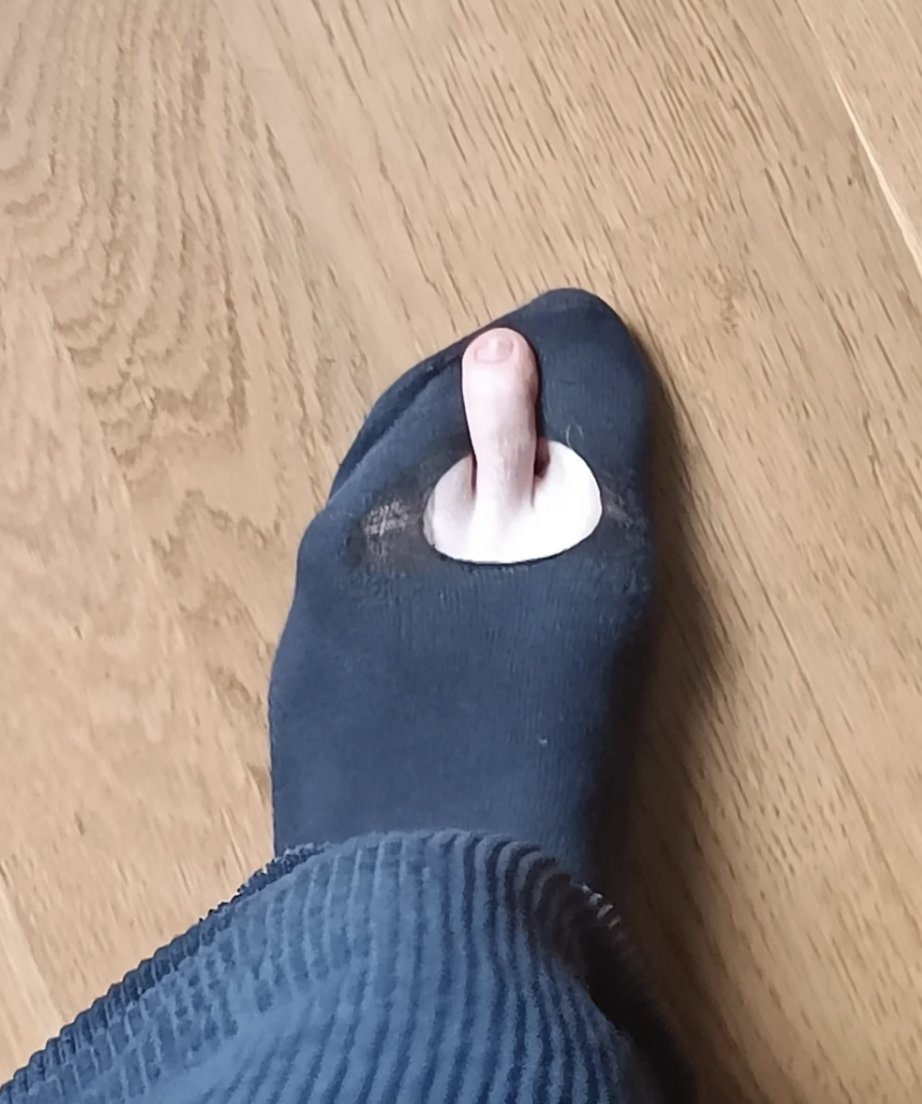 A person's foot in a black sock with a hole, revealing their big toe, standing on a wooden floor