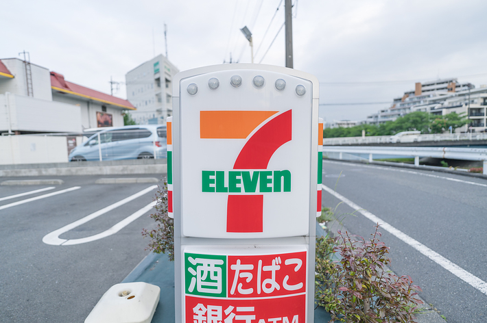 7-Elevenの店舗看板には「酒」「たばこ」「銀行ATM」と書かれている。駐車場と建物が背景に見える。