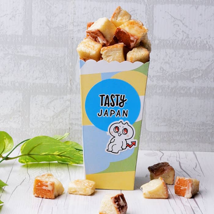 カラフルな箱に入った食パンの一口サイズのキューブが並んでおり、箱には「TASTY JAPAN」と書かれたラベルとマスコットキャラクターが描かれています。