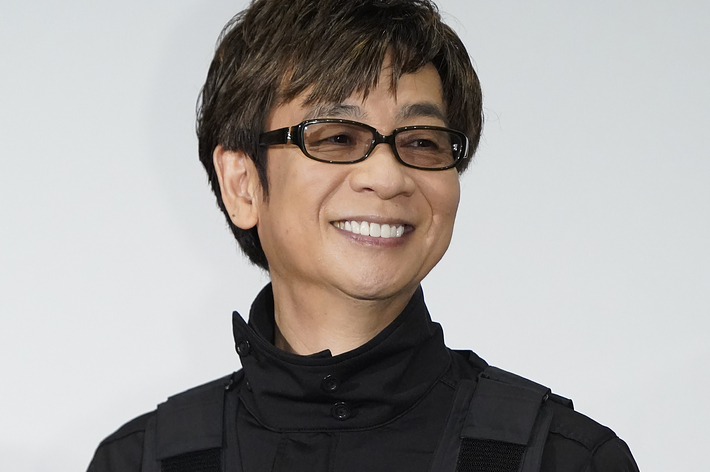 男性がグレーの背景の前で黒いジャケットを着て笑顔で立っています。