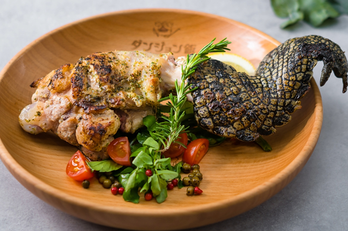 木製の皿にナスの皮で作られた尻尾部分とハーブを添えて焼かれた肉料理が盛り付けられている。