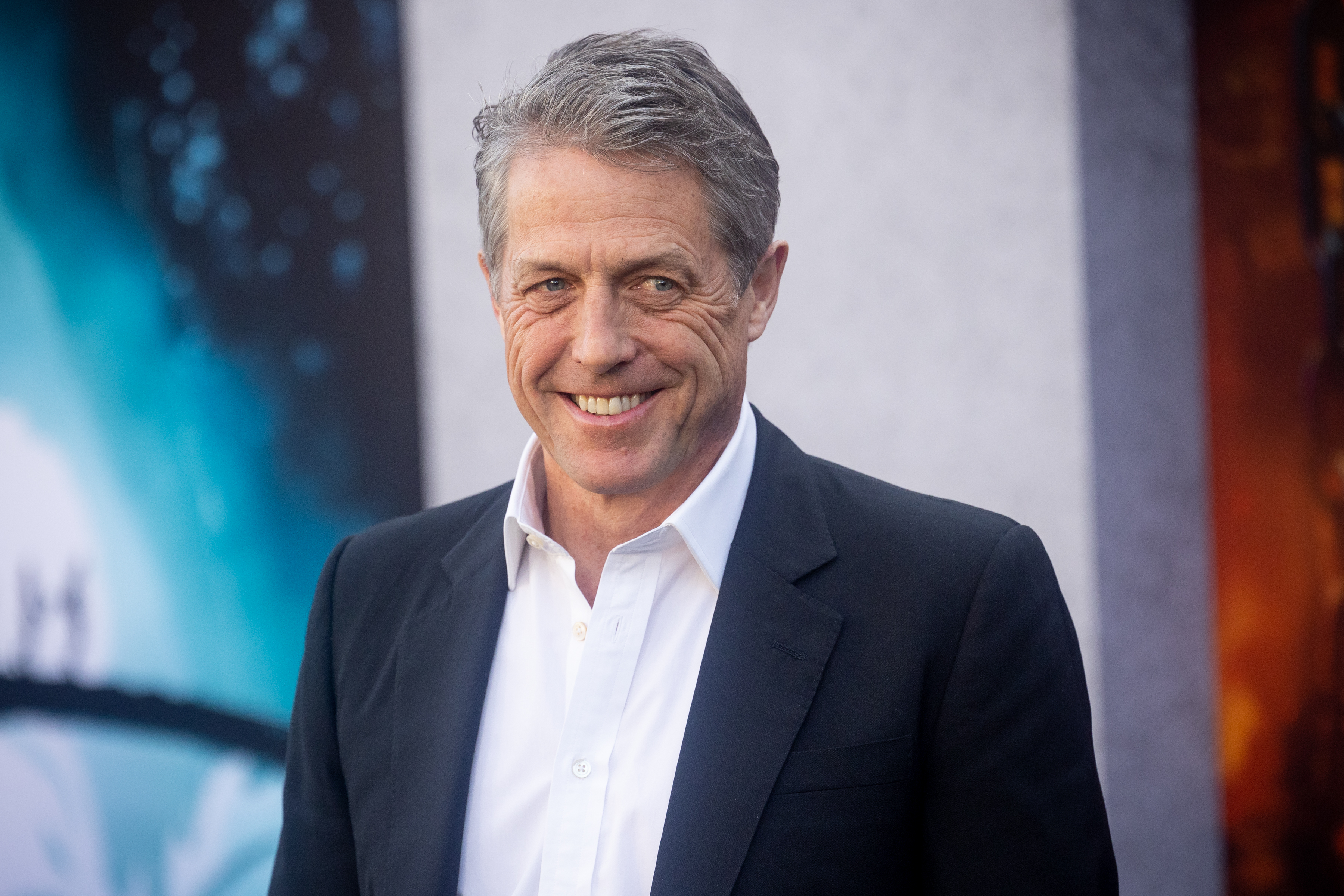 Hugh Grant smiles while wearing a suit at an event
