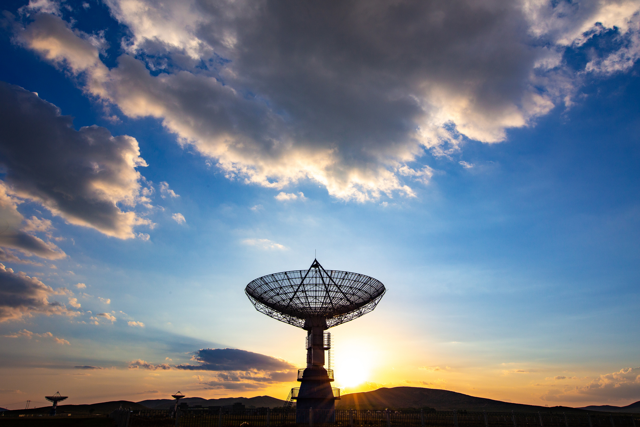 Satellite dish in a field, set against a dramatic sky with clouds and the sun setting behind distant hills