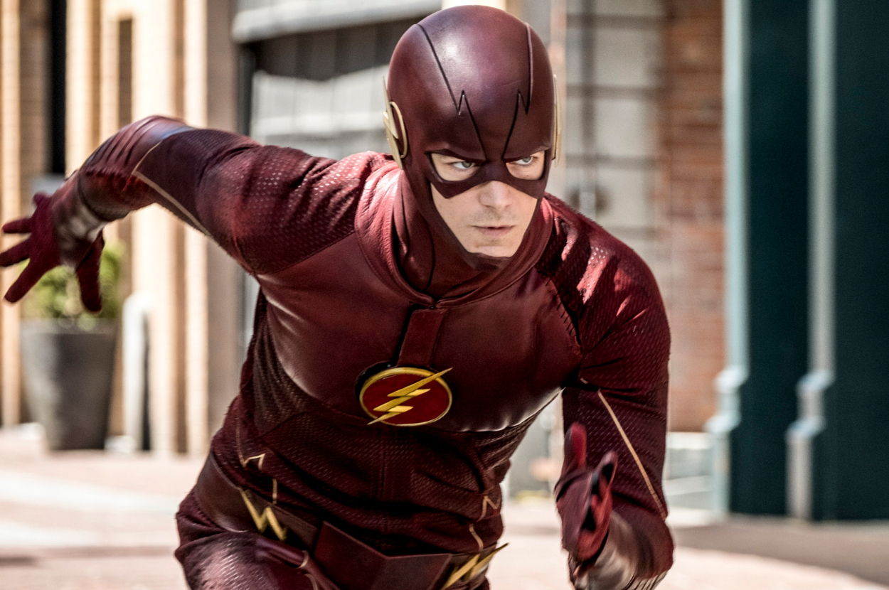 Grant Gustin dressed as The Flash, in a red suit with a lightning bolt emblem, appears to be running fast in an urban setting