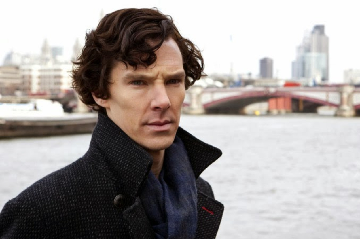 Benedict Cumberbatch stands by a river with a cityscape and bridge in the background, wearing a dark coat and scarf