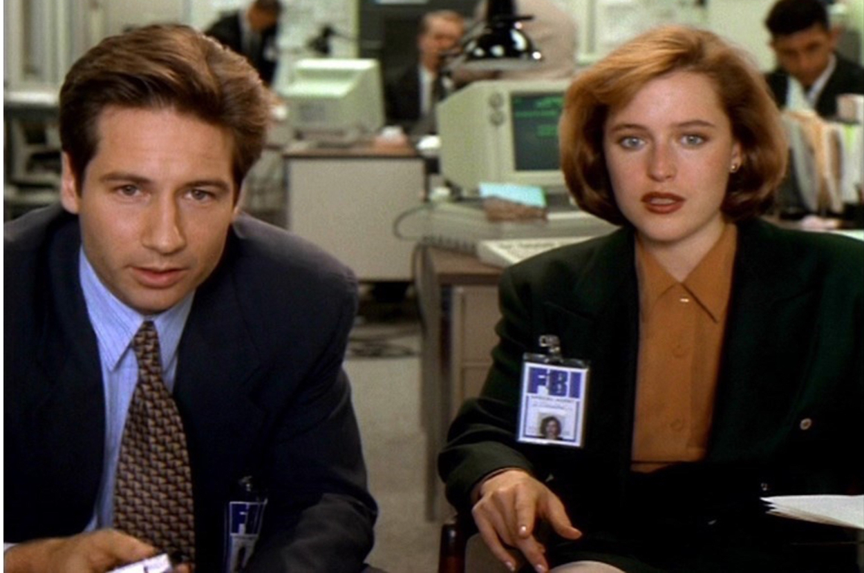 David Duchovny and Gillian Anderson, in office attire, sitting at desks with FBI badges visible, in a scene from The X-Files