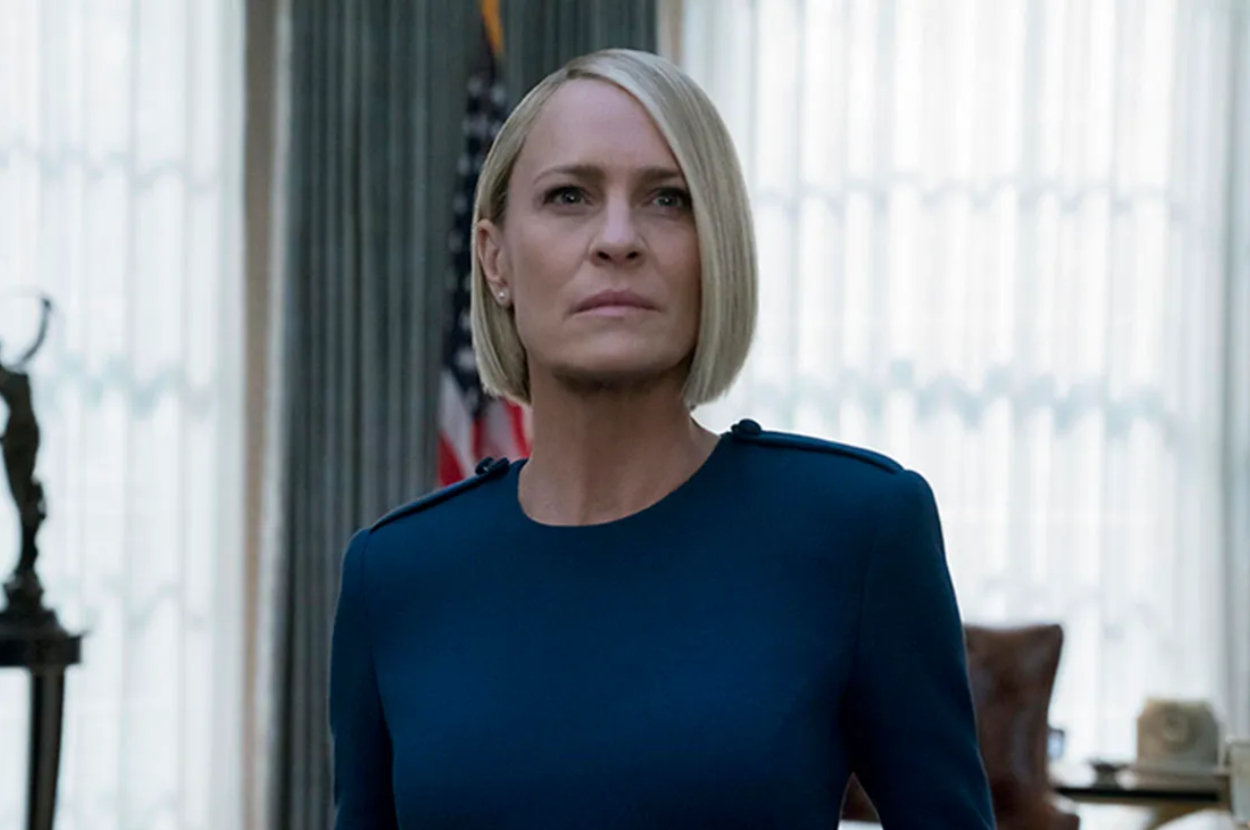 Robin Wright, in a scene, wears a sleek, long-sleeve dress with a minimalist design. An American flag is visible in the background