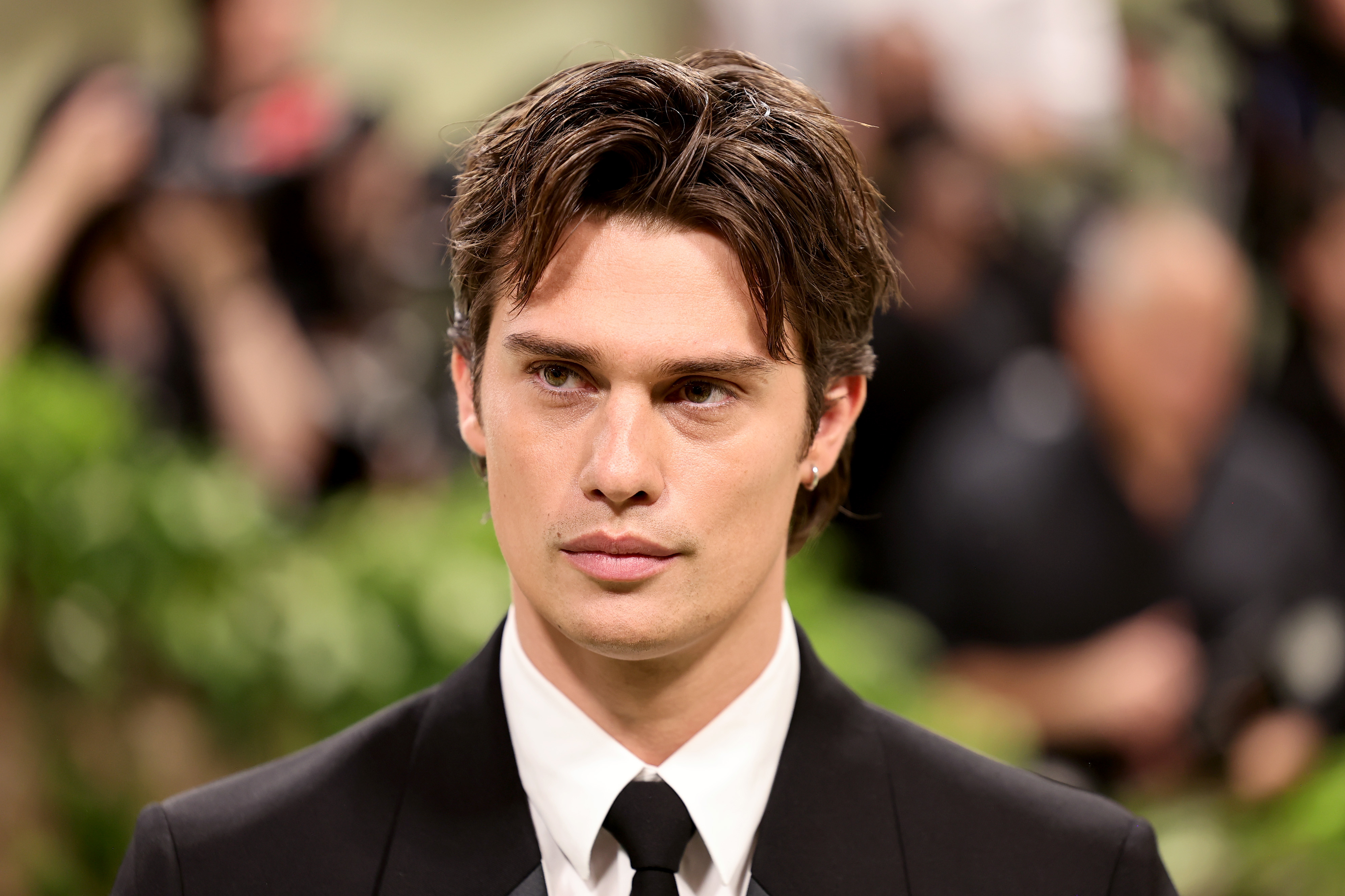 Nicholas Galitzine in a suit and tie poses in a formal setting, with a blurred background of people and greenery