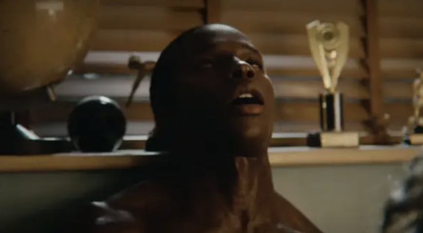 A person is seen sitting with their head tilted back in a room with trophies and a globe in the background