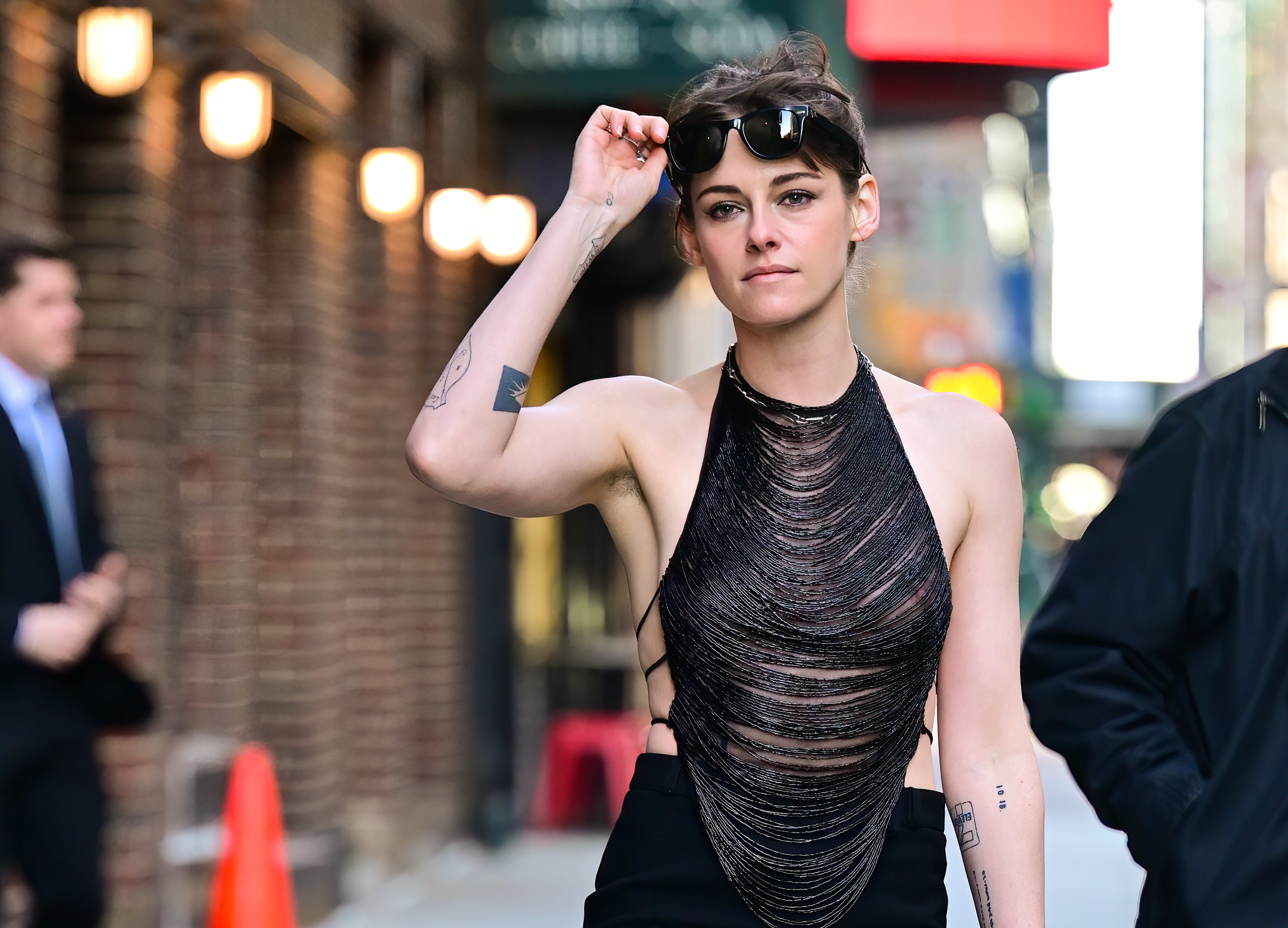 Kristen Stewart walks down a street, lifting sunglasses from her eyes. She wears a sleeveless, open-back, shredded-style top and black pants