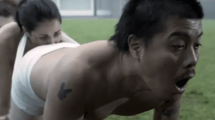 A person with tattoos and short hair is positioned on all fours; another person is behind them. A grassy area and a building are in the background