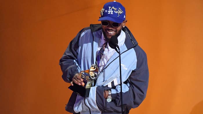 An artist wearing a casual jacket and sunglasses accepts a Grammy award on stage, holding the trophy and smiling