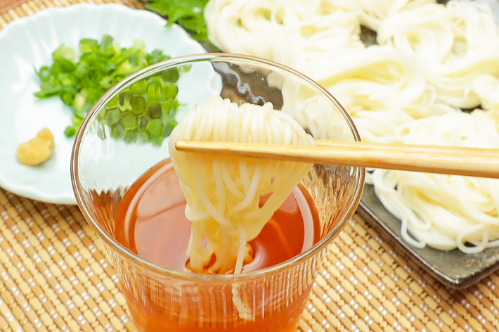 箸でつゆに浸された素麺を持ち上げるシーン。背景には刻んだネギと生姜、お皿に盛られた素麺が写っている。