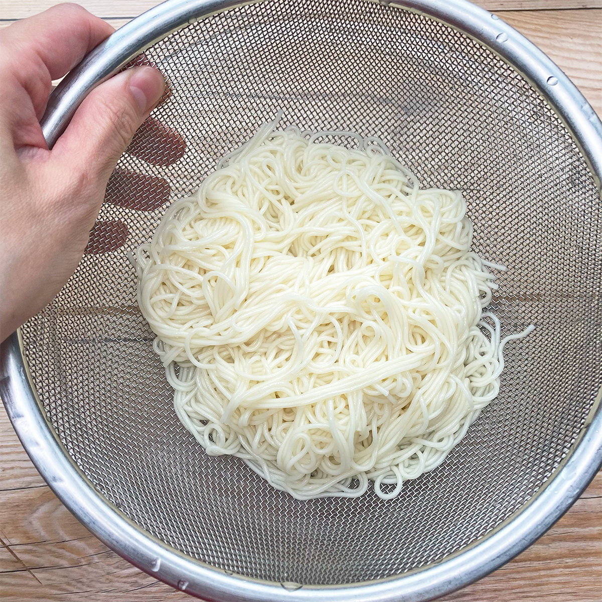 ステンレス製のザルに入ったそうめんを持っている手の写真。