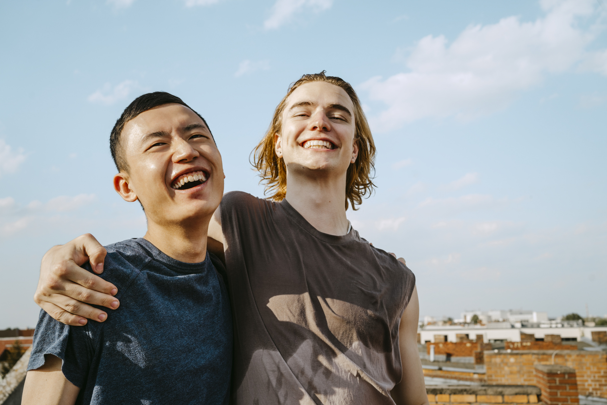 Two people, one with short dark hair and the other with shoulder-length light hair, are standing close together and smiling broadly on a rooftop