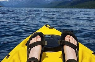 Person's feet in sandals, resting on the front of a yellow kayak on a calm lake with mountains in the background. No visible text or celebrities