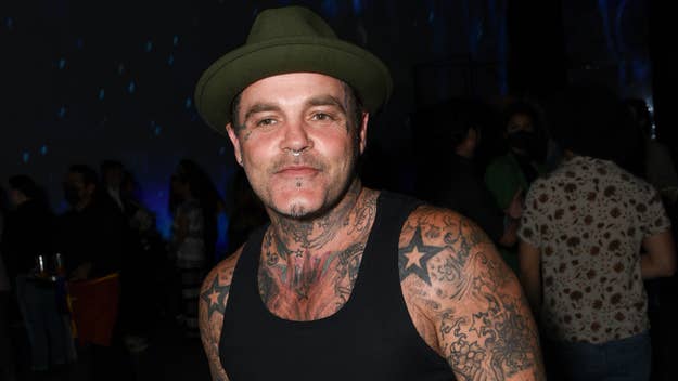Shifty Shellshock with tattooed arms and a tattoo across his neck, wearing a green hat and a black sleeveless shirt, smiles while standing in a crowd at a music event. The name is not provided