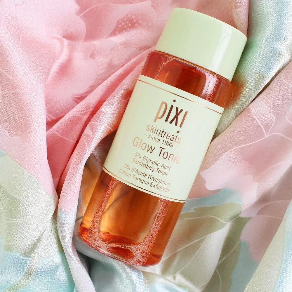 Bottle of Pixi Glow Tonic placed on a pastel patterned fabric backdrop. The label highlights its 5% glycolic acid content and exfoliating properties