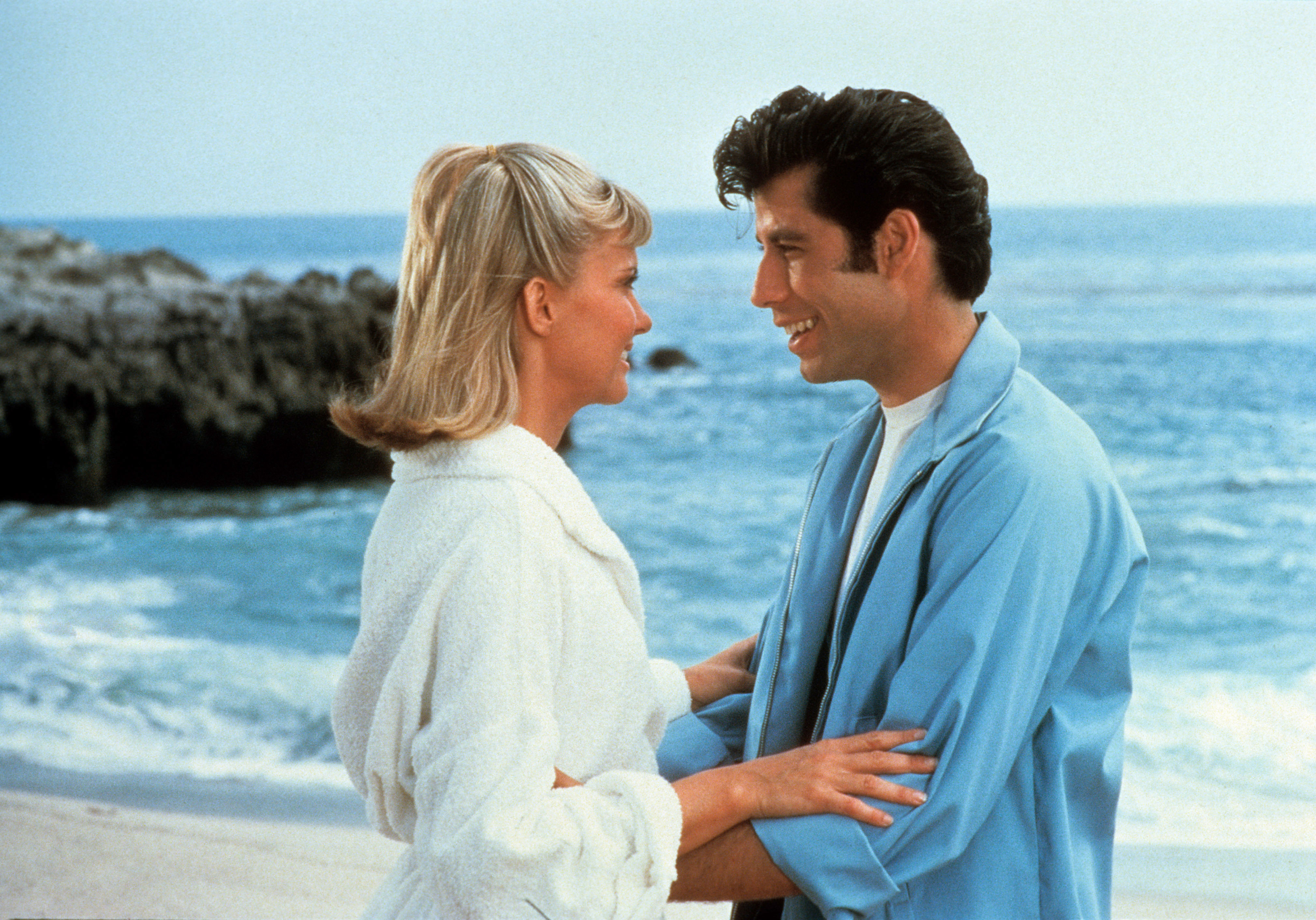 Olivia Newton-John and John Travolta embrace on a beach, smiling at each other, in a scene from the movie "Grease."