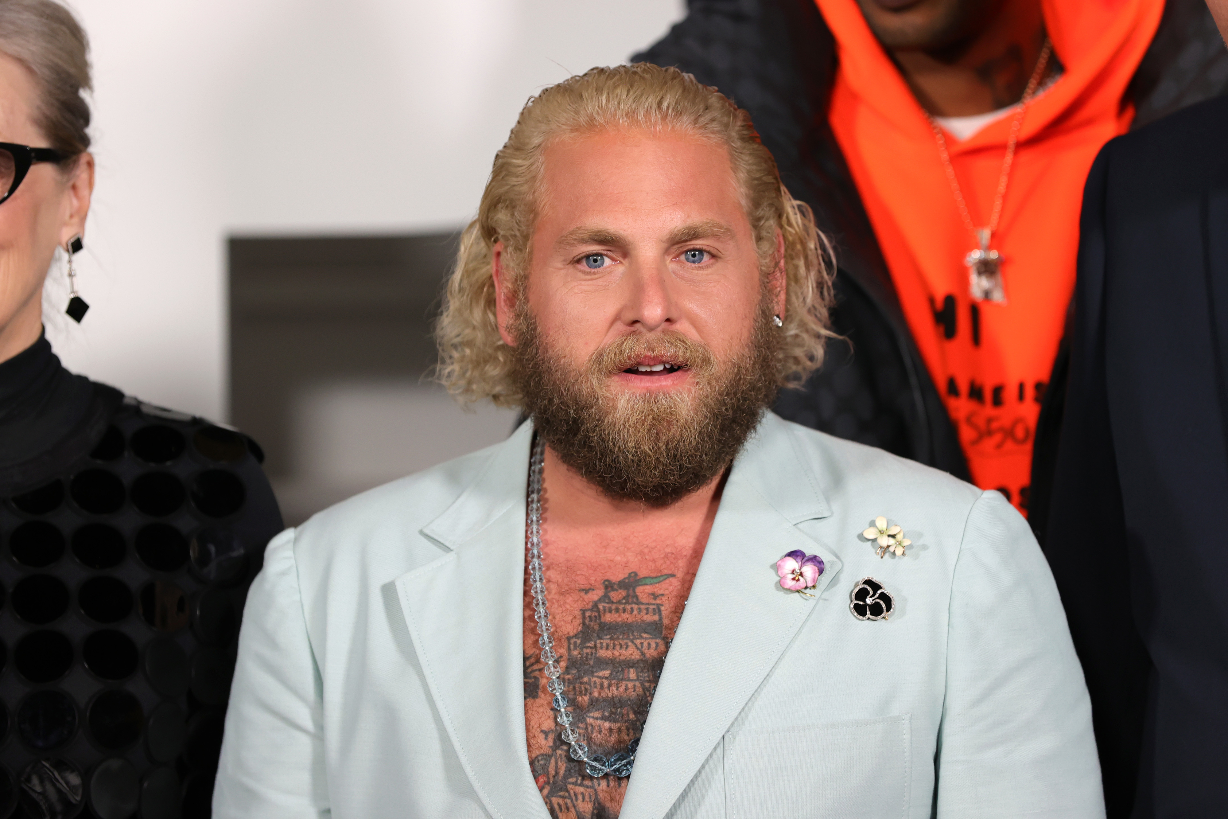 Jonah Hill at a public event wearing a light suit jacket with various pins, his chest tattoos visible