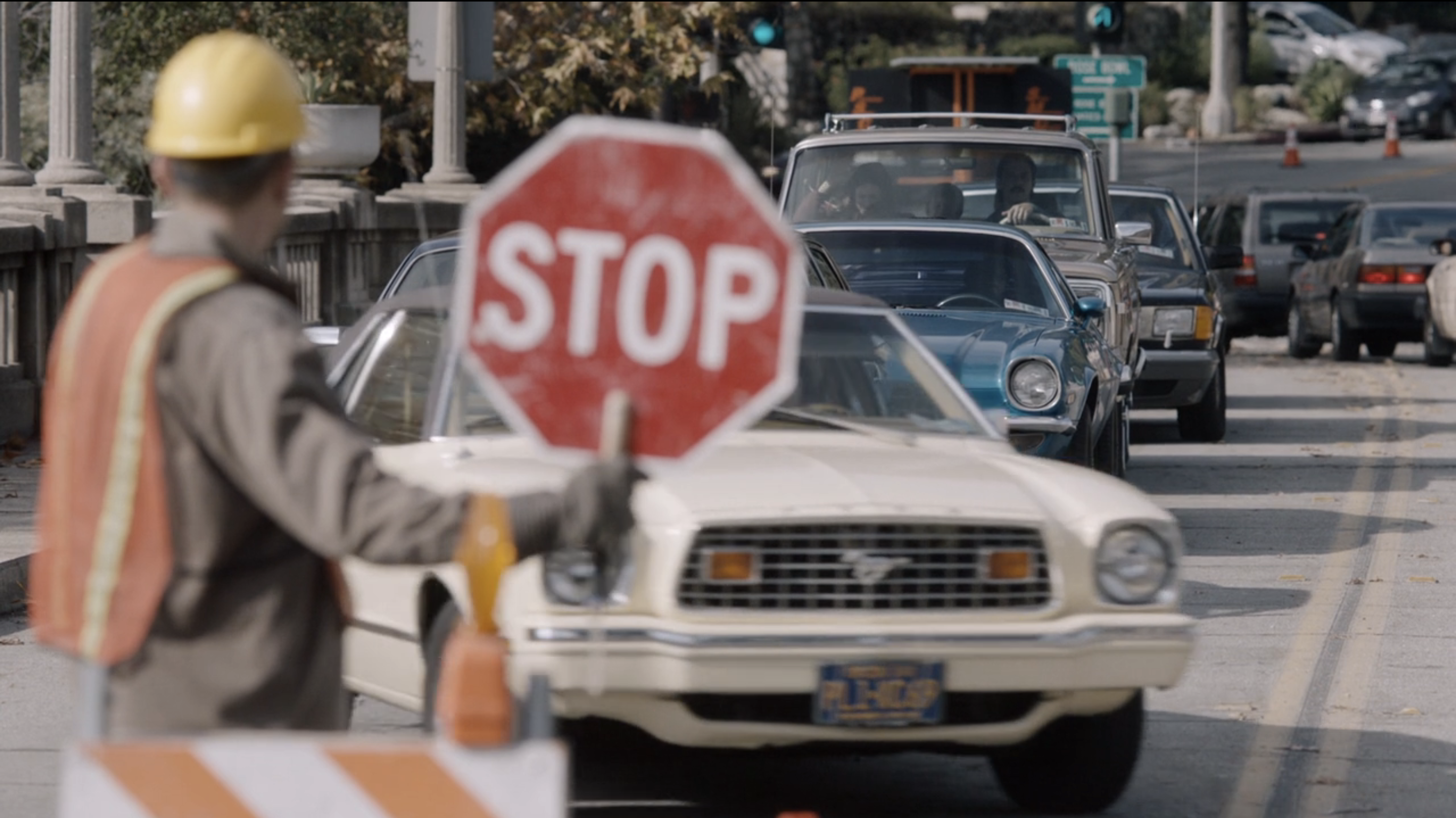 A construction worker holds a "STOP" sign, halting a line of cars on a city street, including a classic Ford Mustang with license plate at the front