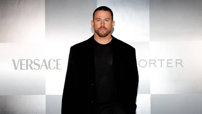 Channing Tatum at a Versace event, wearing a black suit and shirt, posing against a backdrop with Versace and Porter logos