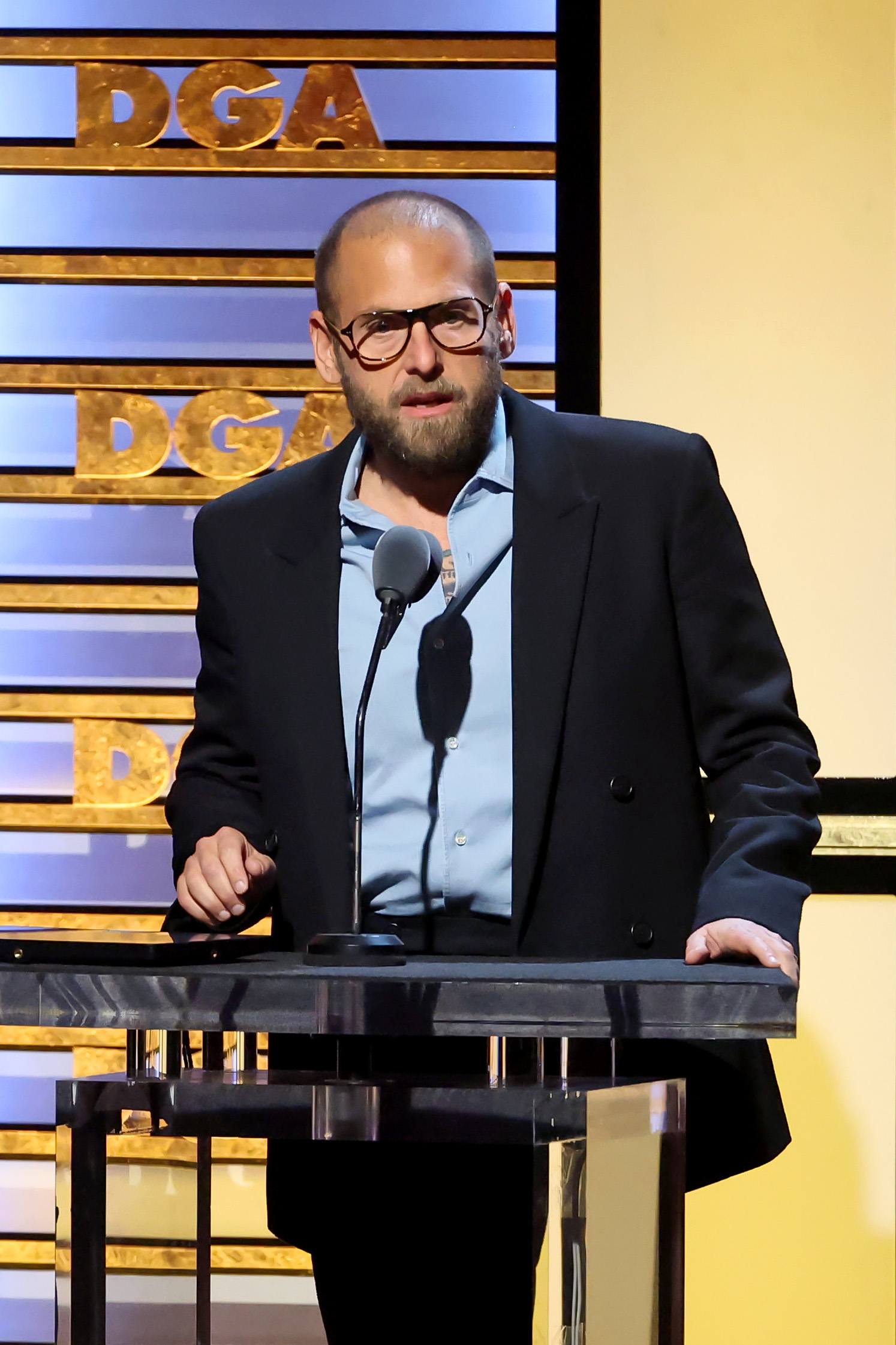 Jonah Hill speaks on stage at the Directors Guild of America awards, wearing a blazer over a light shirt and glasses
