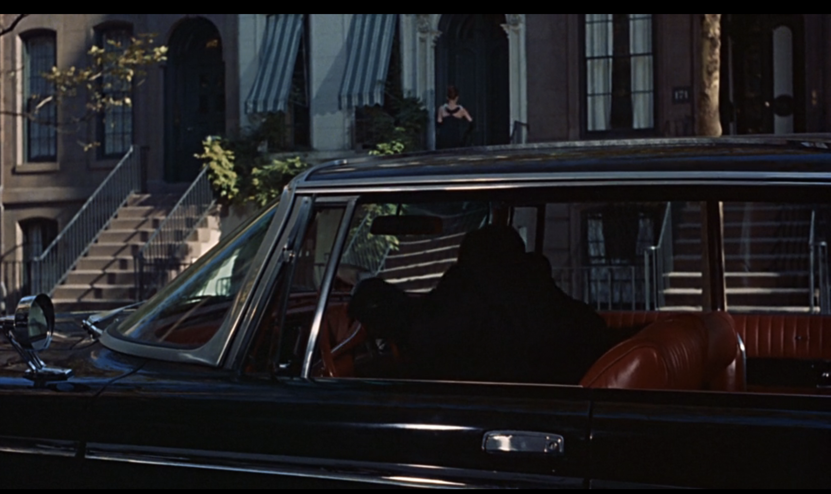 A person dressed in all black sits inside a car parked on a residential street. In the distance, Audrey Hepburn stands on the steps of a building