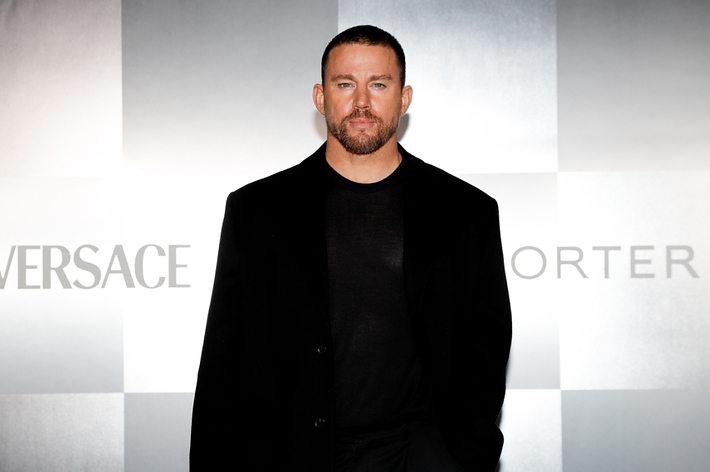Channing Tatum at a Versace event, wearing a black suit and shirt, posing against a backdrop with Versace and Porter logos