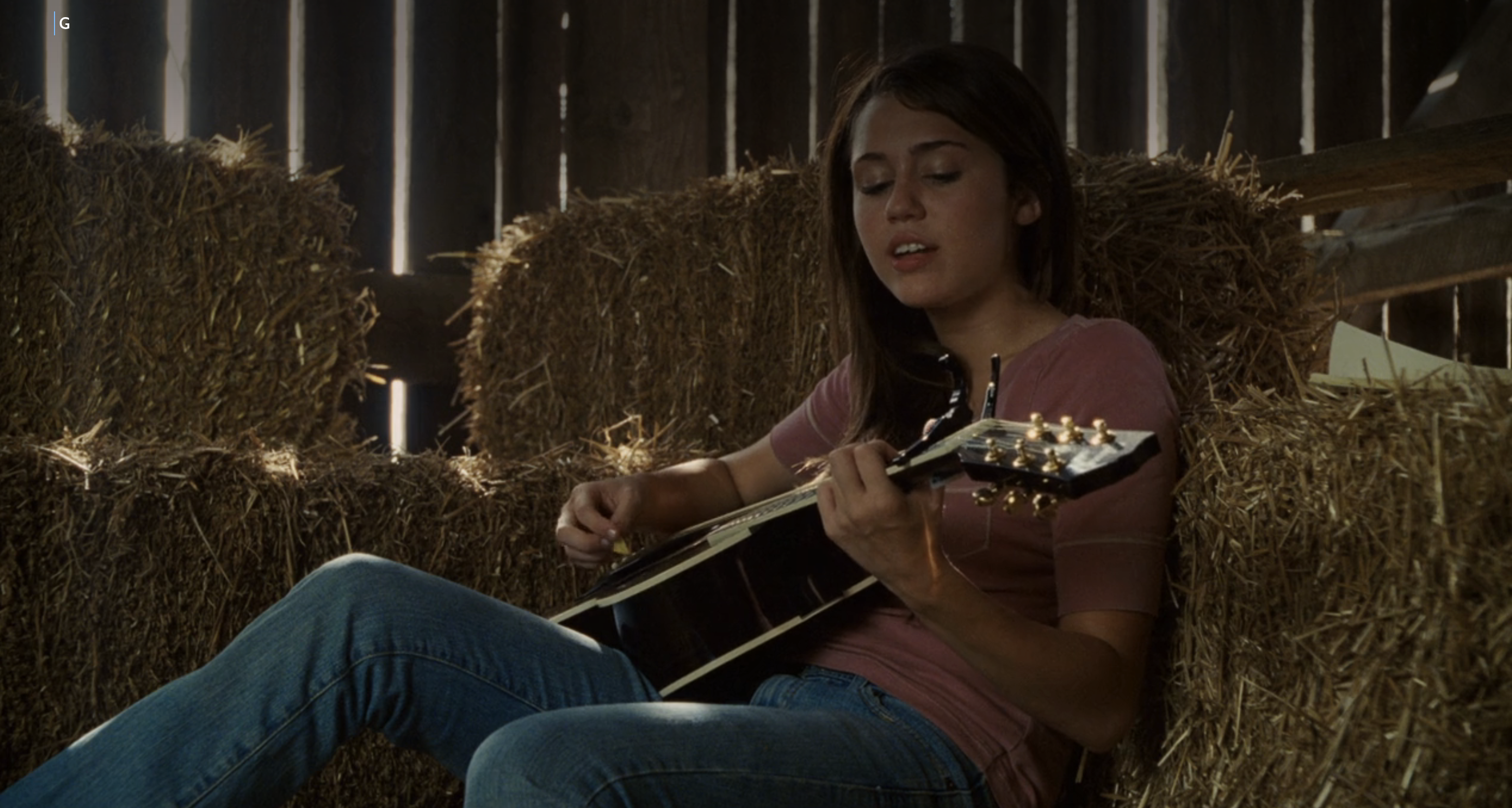 Miley Stewart (Miley Cyrus) plays the guitar while sitting on hay in a barn in a scene from "Hannah Montana: The Movie."