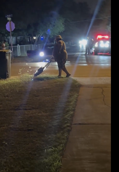 A person is sweeping leaves off the sidewalk at night in a suburban area. Cars with headlights on and one with flashing lights are visible in the background