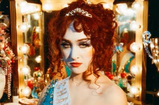 A woman with curly hair, wearing a tiara and a glamorous outfit, stands in front of a vanity mirror with lights