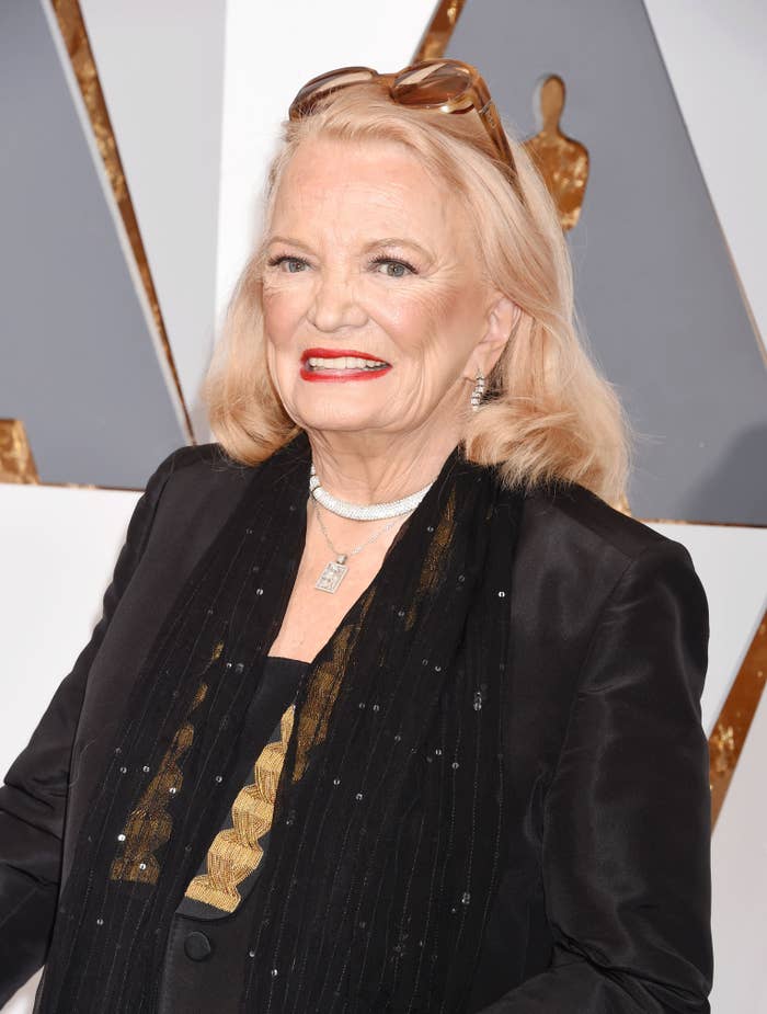 Gena Rowlands on the red carpet wearing a dark outfit with a sheer scarf adorned with gold detailing and beads. She has light hair and is smiling