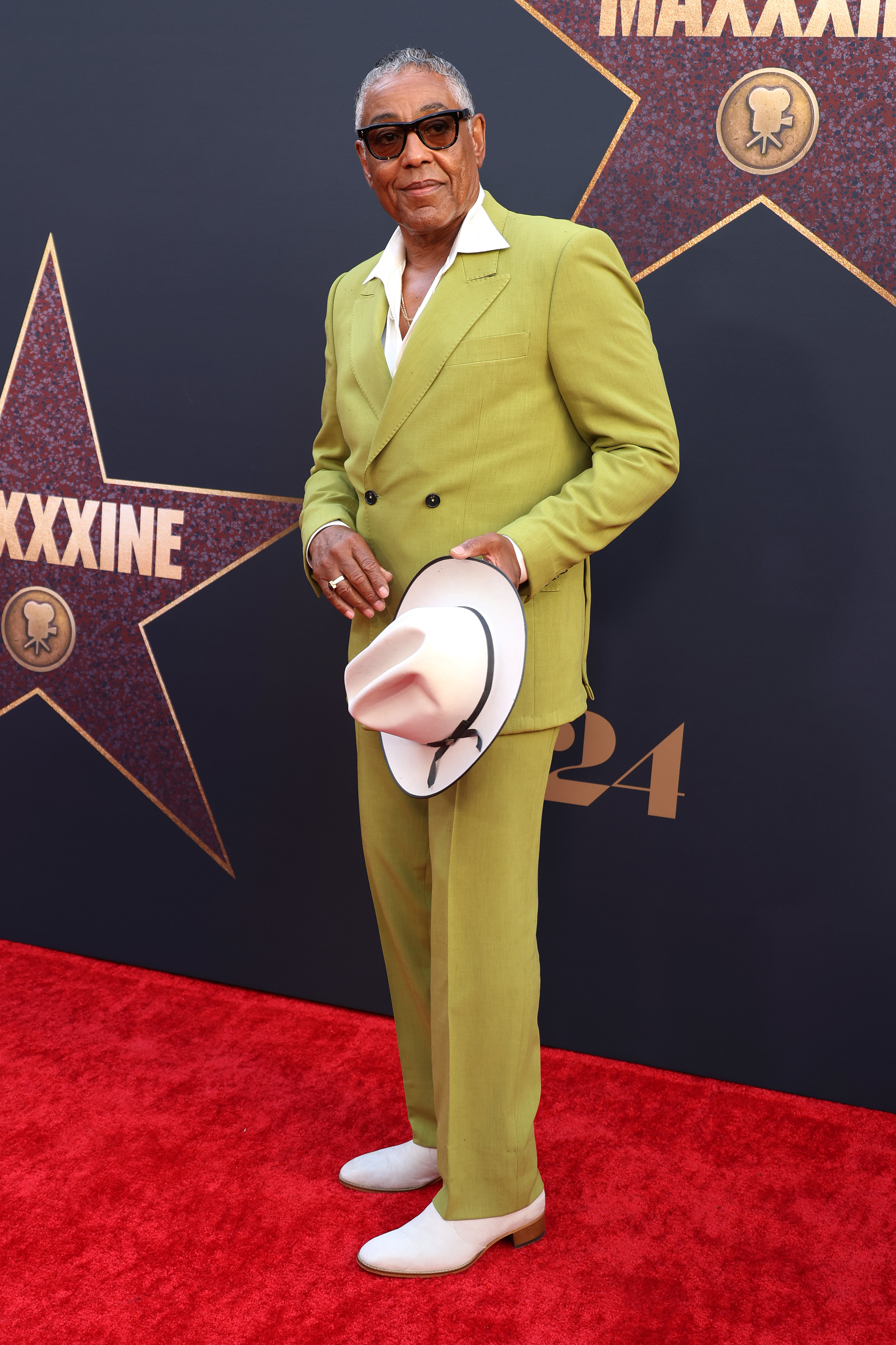 Giancarlo Esposito stands on the red carpet in a stylish suit, holding a fedora hat