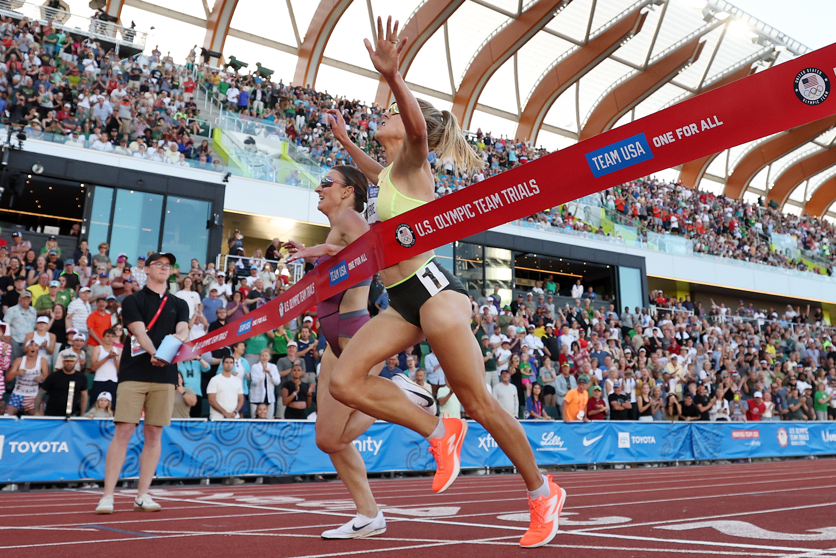 Two female athletes cross the finish line in a race at the U.S. Olympic Team Trials, with a large crowd cheering in the background