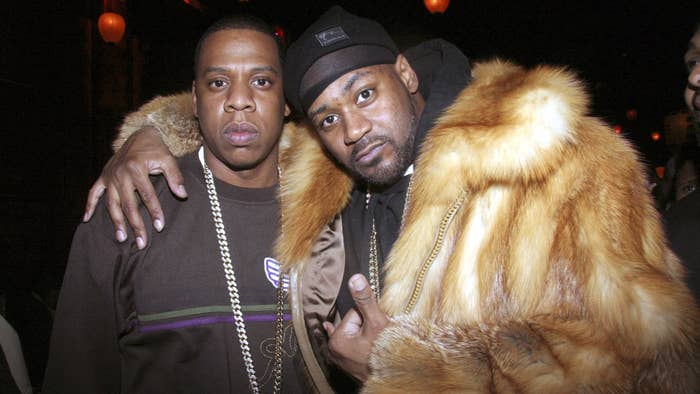Jay-Z and Ghostface Killah pose together. Jay-Z wears a dark sweater with a chain necklace. Ghostface Killah wears a fur coat and a beanie with a chain necklace