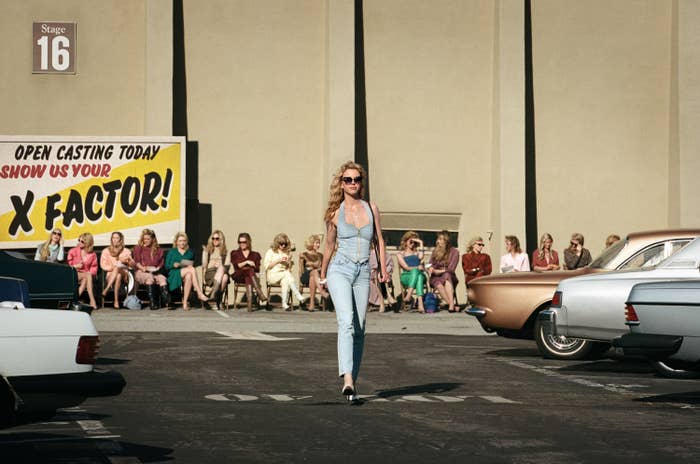 Mia Goth as Maxine walking in a parking lot in a scene from"MaXXXine." There is a row of women sitting behind her for an open casting