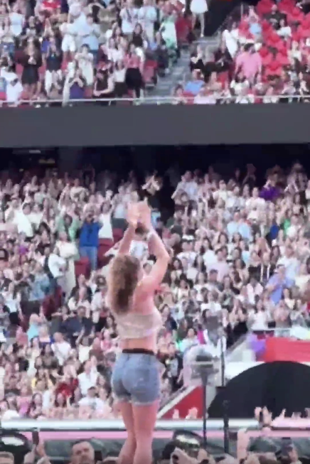 Person on stage facing a cheering stadium crowd with hands raised, wearing jeans and a casual top