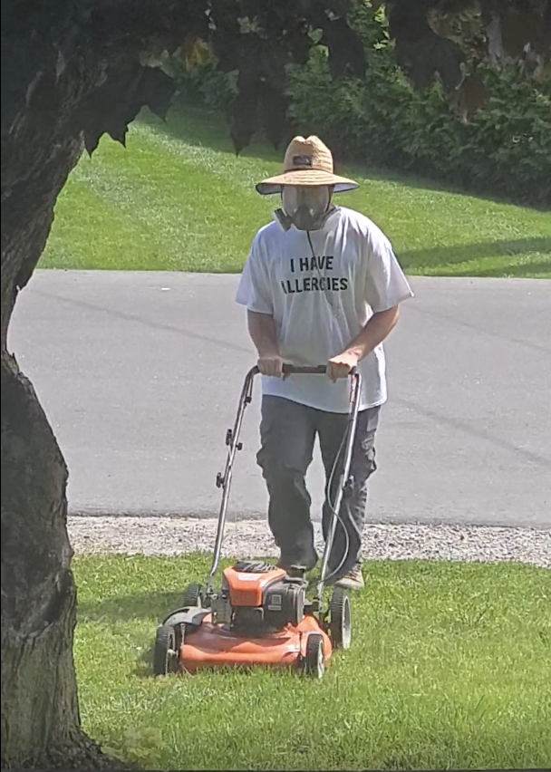 Person mowing the lawn wearing a wide-brimmed hat, a mask, and a shirt that reads &quot;I Have Allergies.&quot; Outdoor scene with greenery in the background