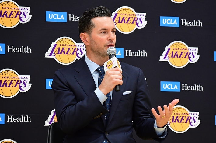 J.J. Redick in a press conference, speaking into a microphone while in a dark suit and tie, seated in front of a Los Angeles Lakers backdrop
