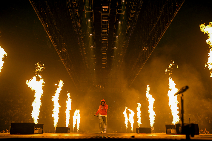 Musician performs on stage amid tall flames and a large crowd at a concert, wearing a hooded sweatshirt