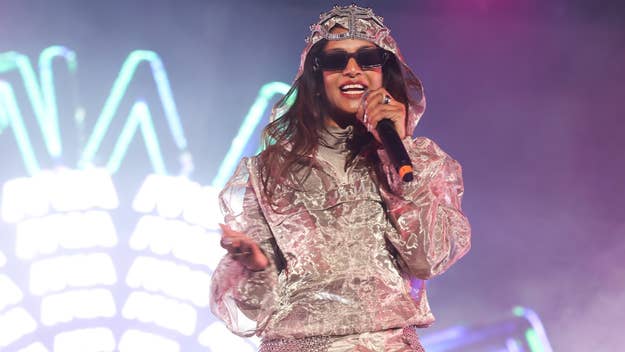 M.I.A. performs on stage in a shiny, metallic outfit with a hood and sunglasses, holding a microphone and smiling. Neon lights are visible in the background