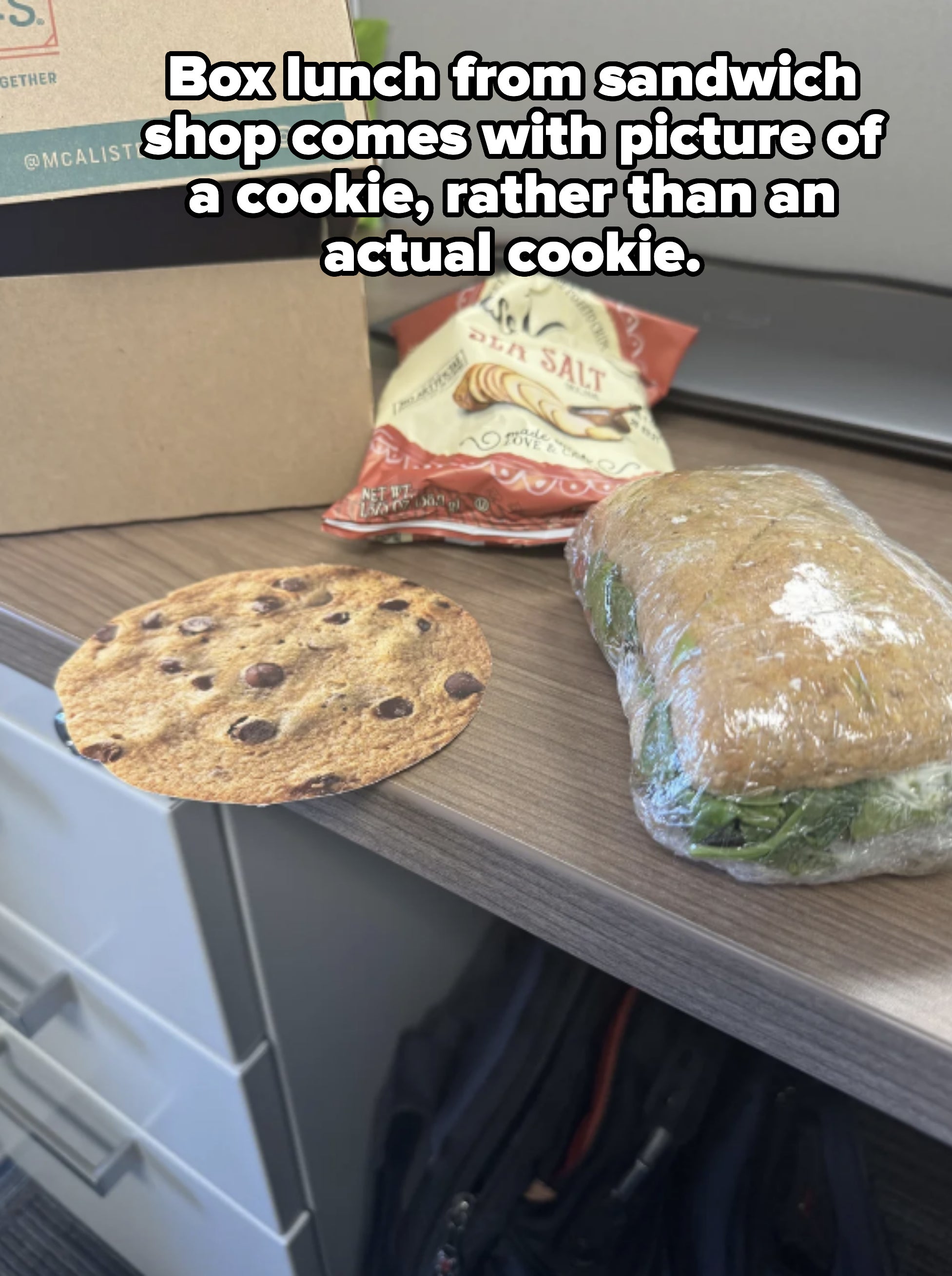 A cookie with chocolate chips, a bag of sea salt chips, and a wrapped sandwich on a desk with a laptop in the background