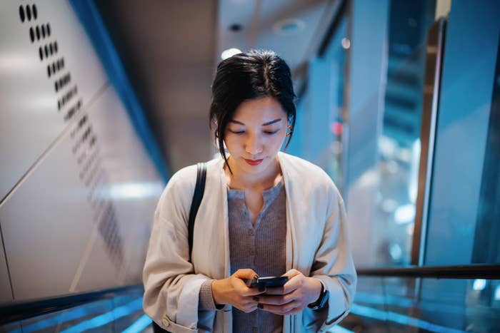 A person in casual attire stands looking at their phone while riding an escalator indoors