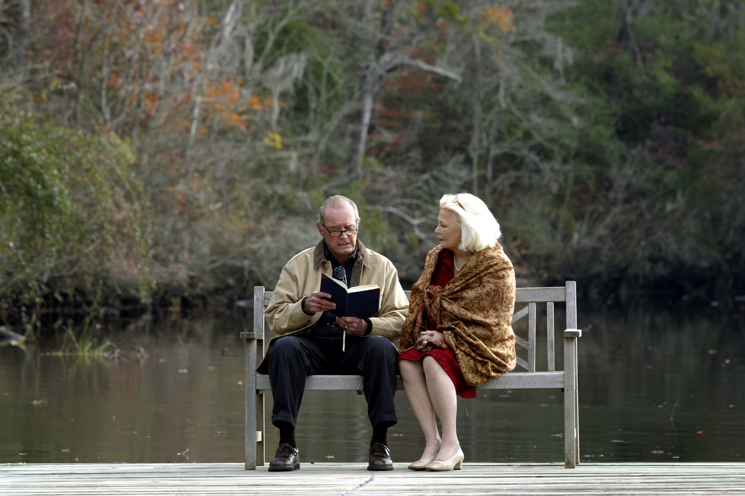James Garner and Gena Rowlands as Noah and Allie in "The Notebook" sitting on a bench in front of a lake. Noah is reading to Allie